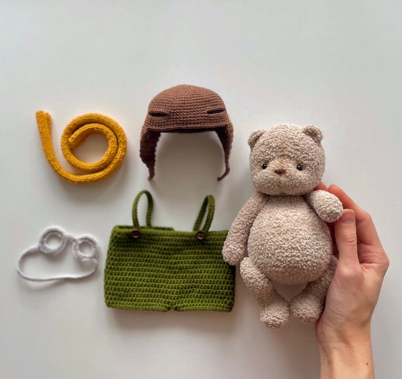 May include: A crocheted teddy bear wearing a brown crocheted hat and green crocheted overalls. The bear is sitting on a white surface.
