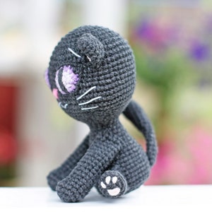 May include: A crocheted black cat toy with purple eyes and a white paw. The cat is sitting on a white surface.