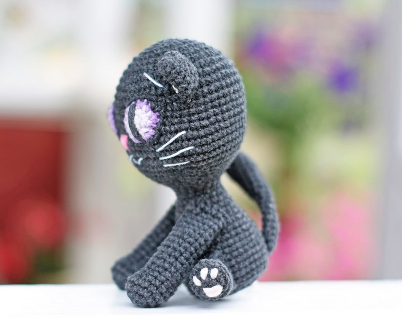 May include: A crocheted black cat toy with purple eyes and a white paw. The cat is sitting on a white surface.