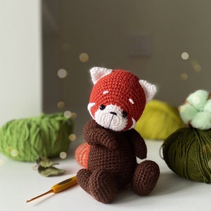 May include: A crocheted red panda plush toy with brown fur and white facial features. The toy is sitting on a white surface with yarn balls in the background.