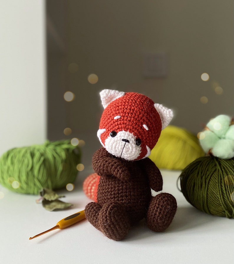 May include: A crocheted red panda plush toy with brown fur and white facial features. The toy is sitting on a white surface with yarn balls in the background.