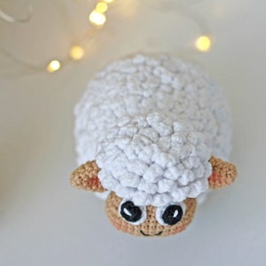 Pu&ograve; includere: Un giocattolo di pecora in crochet bianco con orecchie marroni e occhi neri. La pecora &egrave; seduta su una superficie bianca.