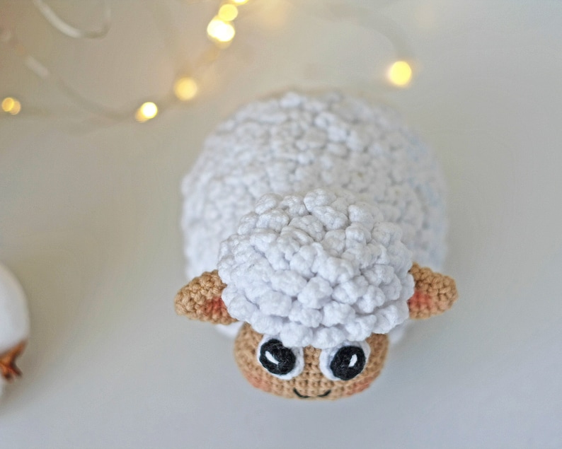 Pu&ograve; includere: Un giocattolo di pecora in crochet bianco con orecchie marroni e occhi neri. La pecora &egrave; seduta su una superficie bianca.