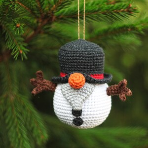 May include: A crocheted snowman ornament with a black top hat, orange nose, and brown arms. The snowman is hanging from a tree branch.