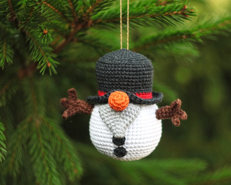 May include: A crocheted snowman ornament with a black top hat, orange nose, and brown arms. The snowman is hanging from a tree branch.