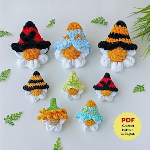 May include: A set of seven crocheted gnome ornaments, each with a different colored hat and a white daisy base. The gnomes are wearing hats in shades of red, blue, yellow, green, and black. The text "PDF Crochet Pattern in English" is visible in the lower right corner of the image.