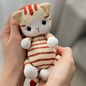 Puede incluir: Un juguete de crochet con forma de gato con rayas blancas y rojas. El gato tiene ojos negros y una nariz blanca.