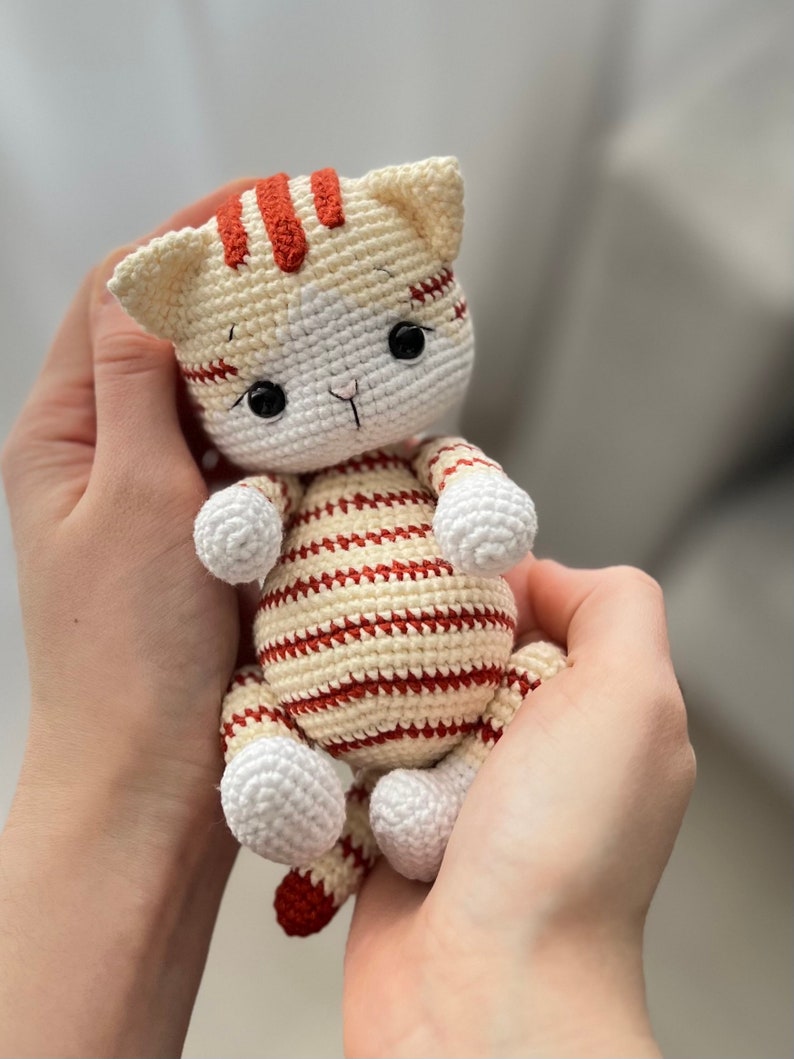 Puede incluir: Un juguete de crochet con forma de gato con rayas blancas y rojas. El gato tiene ojos negros y una nariz blanca.
