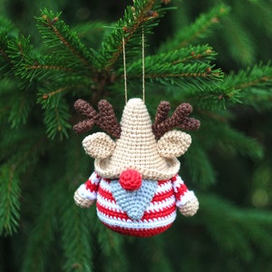 May include: A crocheted reindeer ornament with a red and white striped sweater, brown antlers, and a gray beard. The ornament is hanging from a green pine tree branch.