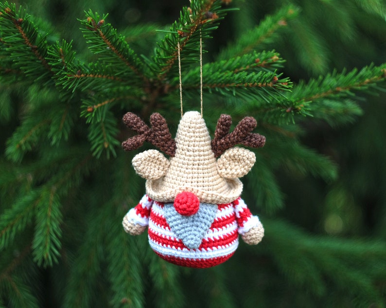 May include: A crocheted reindeer ornament with a red and white striped sweater, brown antlers, and a gray beard. The ornament is hanging from a green pine tree branch.