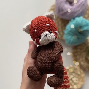 May include: A crocheted red panda plush toy with brown fur and a white face. The toy has black eyes and a black nose.