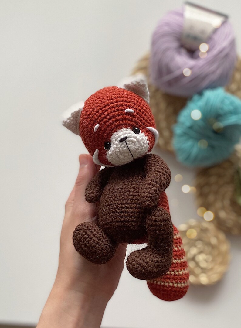 May include: A crocheted red panda plush toy with brown fur and a white face. The toy has black eyes and a black nose.