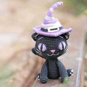 May include: A black crocheted cat wearing a purple and white witch hat with a yellow band. The cat is sitting on a rusty metal surface.