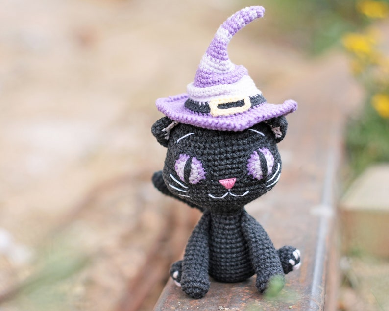 May include: A black crocheted cat wearing a purple and white witch hat with a yellow band. The cat is sitting on a rusty metal surface.