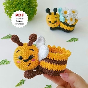 May include: Crocheted bee-themed items. One bee holds a small basket with white daisies. The other bee has a basket-shaped body. The bees are yellow and brown with white wings. The text "PDF Crochet Pattern in English" is in a white circle.