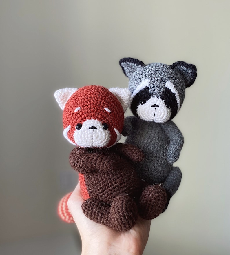 May include: A crocheted red panda plush toy with brown fur and white facial features, sitting on a hand. Behind the red panda is a crocheted gray raccoon plush toy with black eyes and a white nose.
