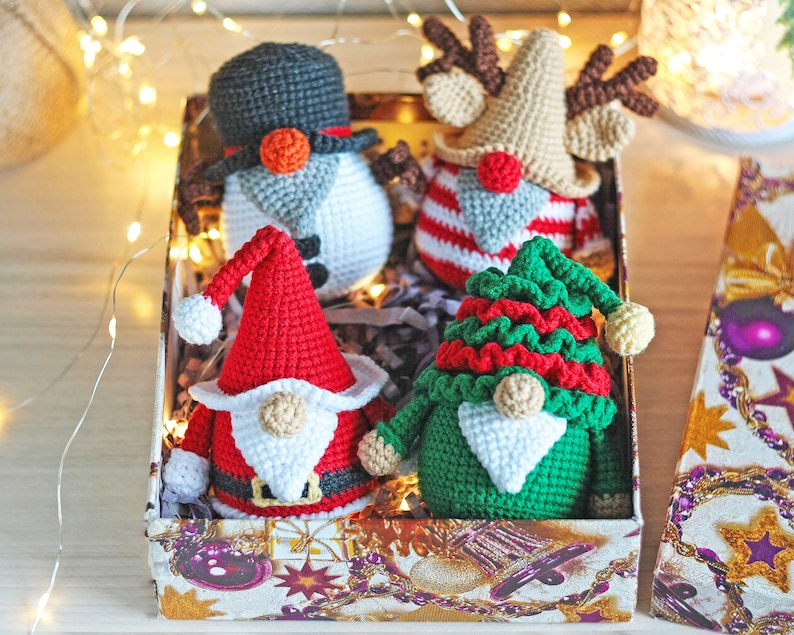 May include: A set of four crocheted Christmas gnomes in a gift box. The gnomes are dressed in red, green, white, and brown. The box is decorated with a Christmas theme.