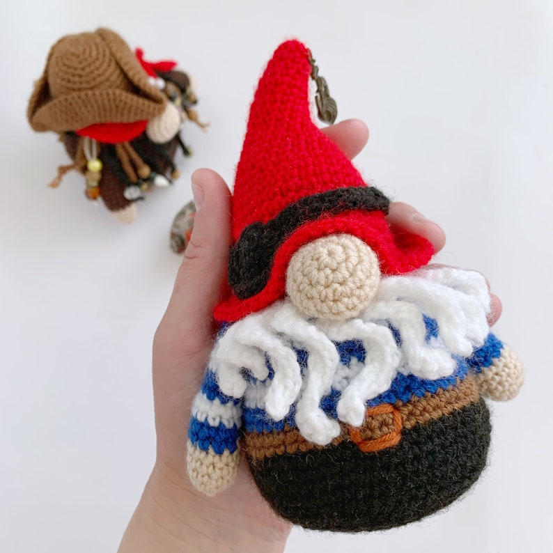 May include: A crocheted gnome wearing a red hat with a black eye patch, a blue and white striped shirt, and a brown belt. The gnome has a white beard and a brown crocheted body.