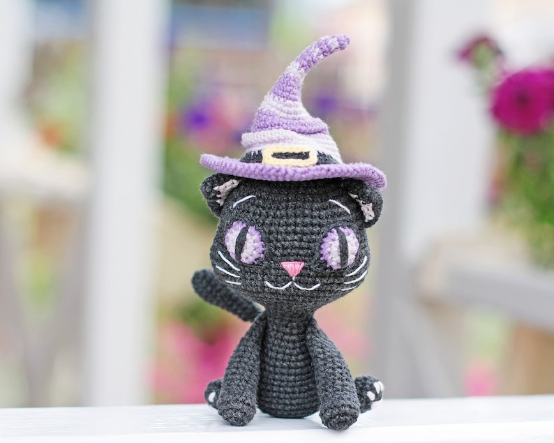 May include: A black crocheted cat wearing a purple and white witch hat. The cat has large, white eyes and a pink nose.