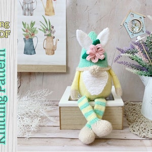 Puede incluir: Patrón de tejido para un gnomo amarillo y verde con barba blanca y una flor rosa en su sombrero. El gnomo está sentado en una caja de madera con un borde blanco. El patrón está disponible en inglés y es una descarga PDF.