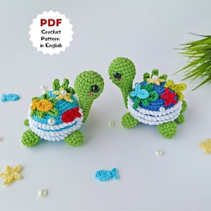 May include: Two crocheted green sea turtles with colorful shells. The shells are decorated with flowers, starfish, and fish. The turtles are sitting on a white surface with white pearls scattered around them.
