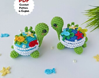 May include: Two crocheted green sea turtles with colorful shells. The shells are decorated with flowers, starfish, and fish. The turtles are sitting on a white surface with white pearls scattered around them.