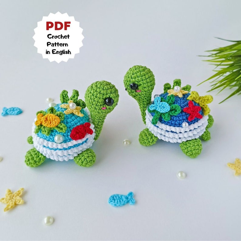 May include: Two crocheted green sea turtles with colorful shells. The shells are decorated with flowers, starfish, and fish. The turtles are sitting on a white surface with white pearls scattered around them.