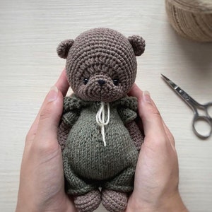 May include: A crocheted brown bear wearing a green crocheted outfit. The bear is being held by two hands.