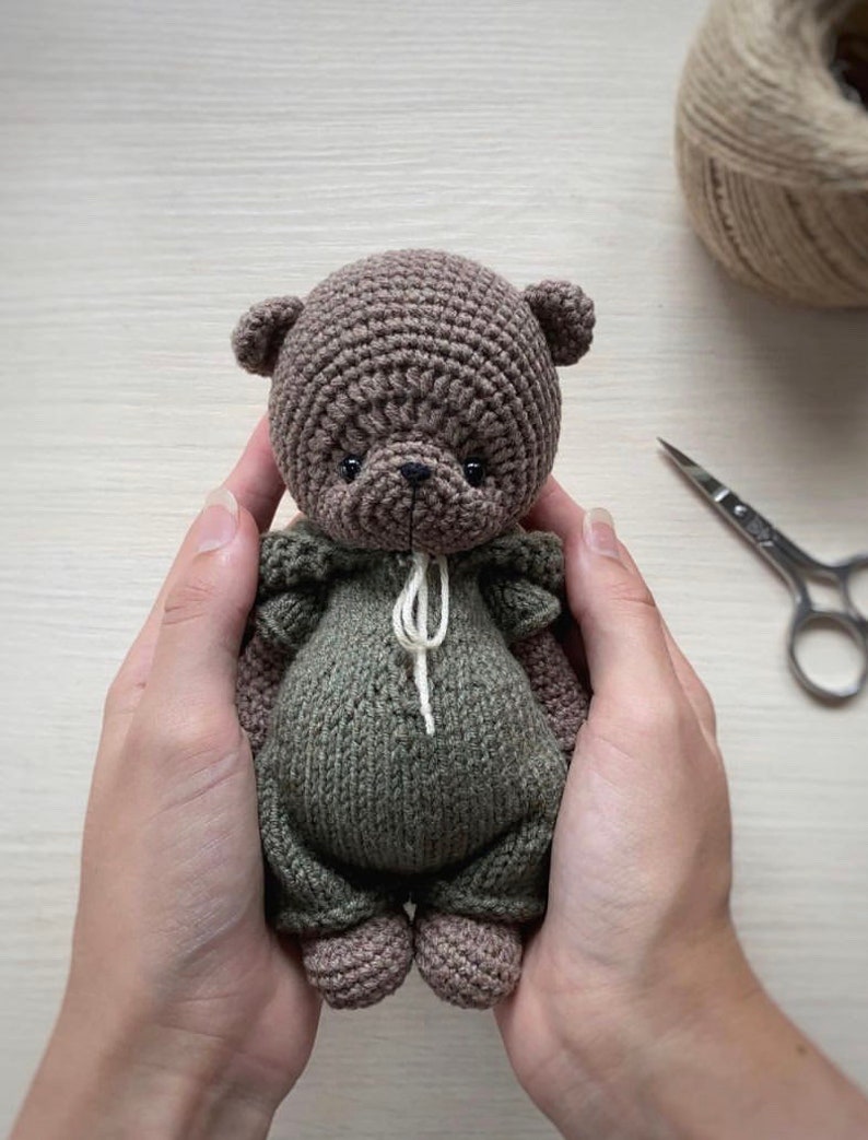 May include: A crocheted brown bear wearing a green crocheted outfit. The bear is being held by two hands.
