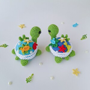 May include: Two crocheted green sea turtles with colorful flowers, starfish, and fish on their shells. The turtles are facing each other and have white pearl accents.