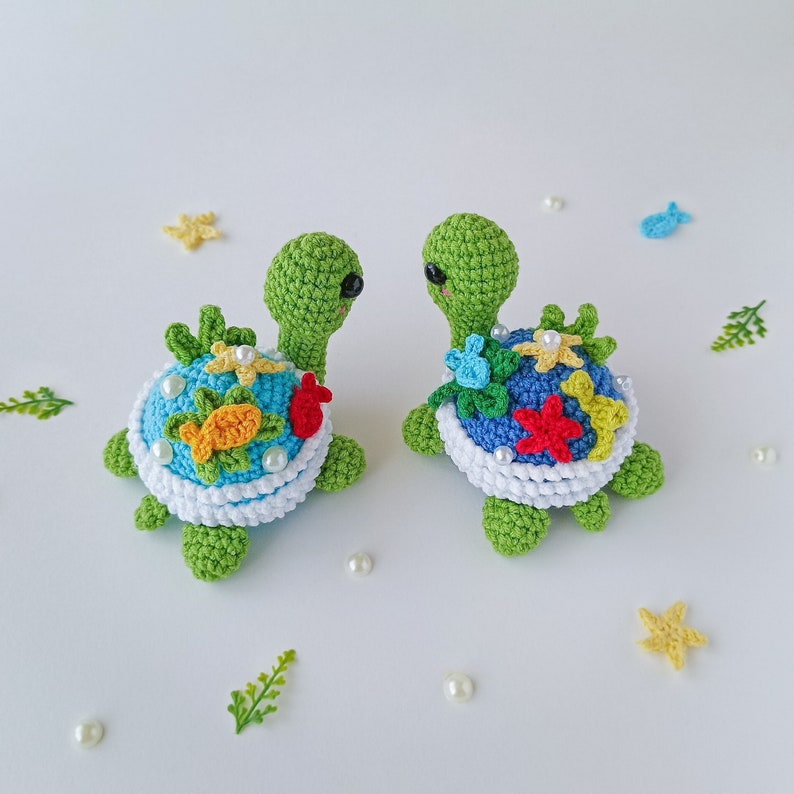 May include: Two crocheted green sea turtles with colorful flowers, starfish, and fish on their shells. The turtles are facing each other and have white pearl accents.