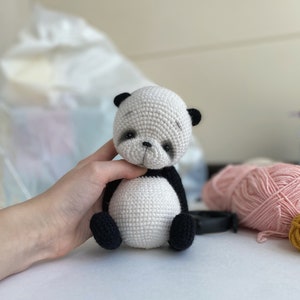 May include: A small crocheted panda bear toy with black ears, eyes, and paws. The panda is sitting on a white surface.