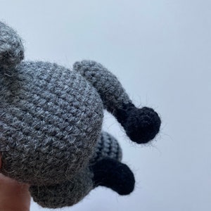 May include: A crocheted raccoon plush toy with gray fur and black feet. The toy is being held by a hand.