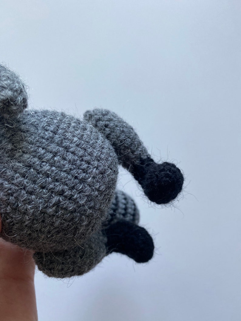 May include: A crocheted raccoon plush toy with gray fur and black feet. The toy is being held by a hand.