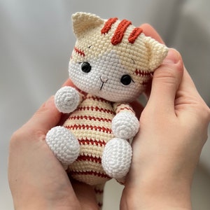 Puede incluir: Un juguete de crochet con forma de gato con rayas blancas y rojas. El gato tiene ojos negros y una cara blanca.