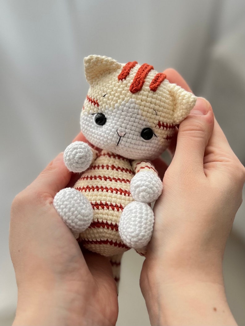 Puede incluir: Un juguete de crochet con forma de gato con rayas blancas y rojas. El gato tiene ojos negros y una cara blanca.
