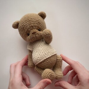 May include: A crocheted brown teddy bear wearing a white knitted shirt. The bear is being held by two hands.