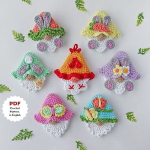 Peut inclure: Un ensemble de 7 gnomes en crochet avec des chapeaux et des décorations de différentes couleurs. Les gnomes ont des barbes blanches et des joues roses. Les chapeaux sont décorés de papillons, de fleurs et de lapins. Les gnomes sont disposés sur un fond blanc avec des feuilles vertes.