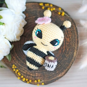 May include: A crocheted bee toy with black and yellow stripes, holding a small jar of honey that says "Honey". The bee has a pink flower on its head and large blue eyes.