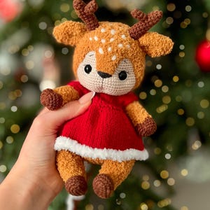 May include: A handmade crochet deer doll with a red dress and white trim. The deer has brown antlers, ears, and hooves, with white spots on its head. The doll is a soft, plush toy.