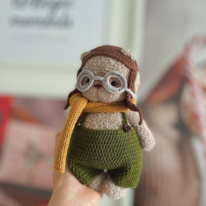 May include: A crocheted stuffed animal wearing a brown aviator hat, round white glasses, and a yellow scarf. The animal is wearing green overalls with a brown button.