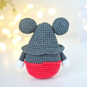 May include: A crocheted stuffed toy resembling Mickey Mouse. The toy is gray with red shorts and white gloves. The toy is standing on a white surface.
