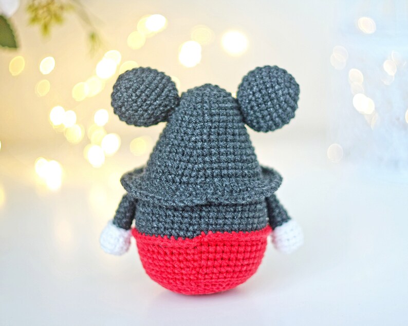 May include: A crocheted stuffed toy resembling Mickey Mouse. The toy is gray with red shorts and white gloves. The toy is standing on a white surface.