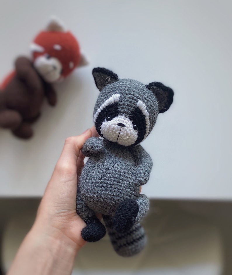 May include: A crocheted raccoon plush toy with a gray body, black eyes, white nose, and black and white striped tail.