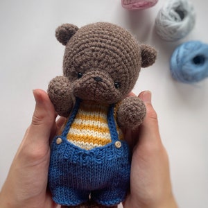 May include: A brown crocheted teddy bear wearing a blue and yellow striped knitted outfit. The bear is being held by a person's hands.