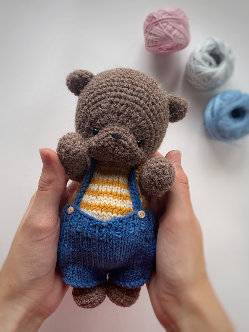 May include: A brown crocheted teddy bear wearing a blue and yellow striped knitted outfit. The bear is being held by a person's hands.