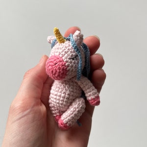 May include: A small pink crocheted unicorn with a yellow horn and blue mane. The unicorn is lying on its side with its eyes closed.