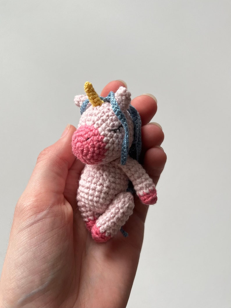 May include: A small pink crocheted unicorn with a yellow horn and blue mane. The unicorn is lying on its side with its eyes closed.