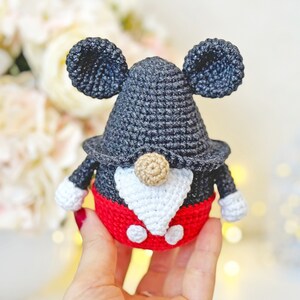 May include: A crocheted gnome with black and white ears, a black hat, a white beard, and a red and white outfit. The gnome is wearing a red and white outfit with black buttons.