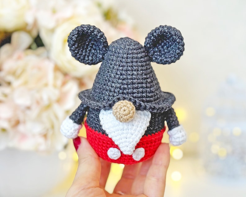 May include: A crocheted gnome with black and white ears, a black hat, a white beard, and a red and white outfit. The gnome is wearing a red and white outfit with black buttons.
